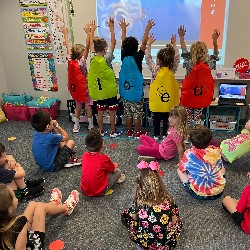 Five students dressed in capes with vowels printed on the back of each cape.